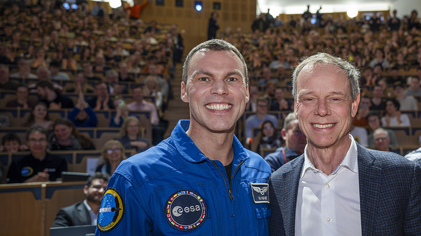 Två män ler med en stor publik i en hörsal i bakgrunden.