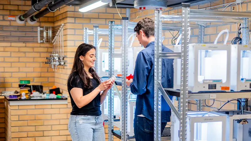 Två studenter som står bredvid hyllor fyllda med 3D-skrivare.